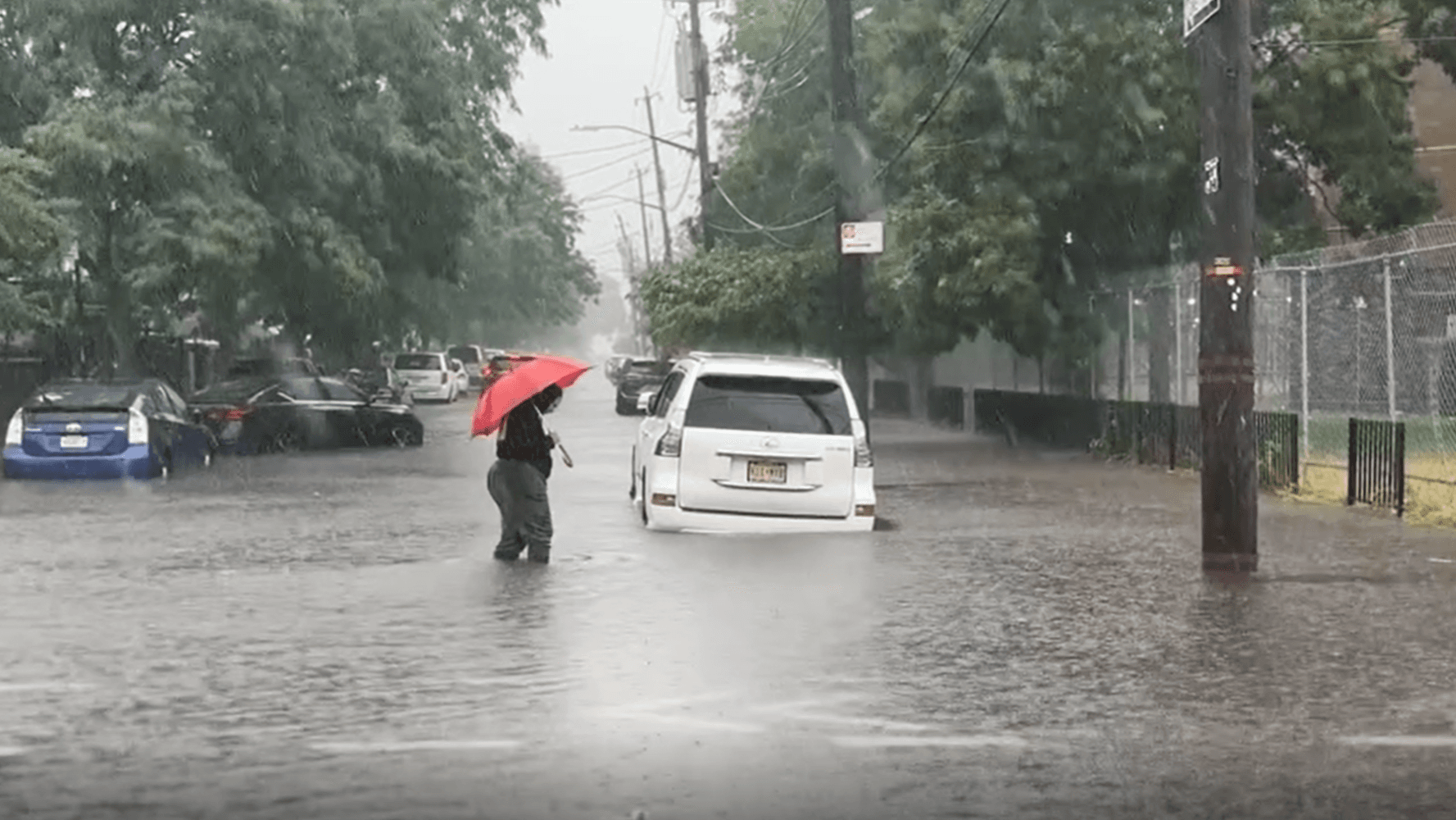 A person carrying a red umbrella crosses a flooded street in Brooklyn, up to their knees in stormwater